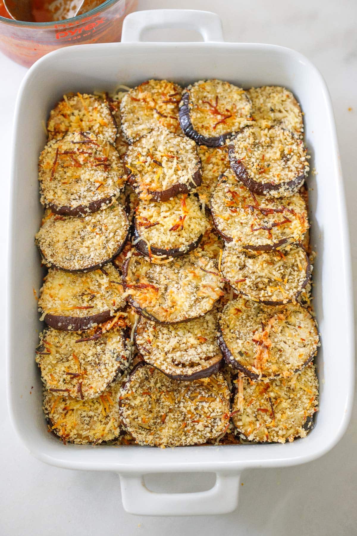 white baking dish with layer of breaded eggplant slices with crispy, golden parmesan